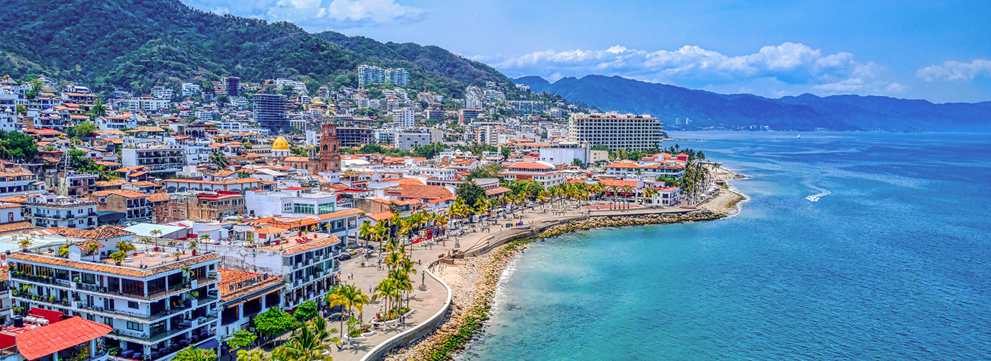 Vista aérea del malecón de Puerto Vallarta, mostrando el paseo marítimo, la playa, y la vibrante zona urbana rodeada de montañas y el océano Pacífico.