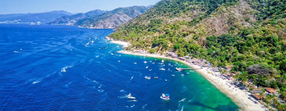 Vista aérea panorámica de Playa Las Ánimas en Puerto Vallarta, mostrando la costa con arena clara, agua turquesa, vegetación exuberante en las montañas y botes anclados en la bahía.